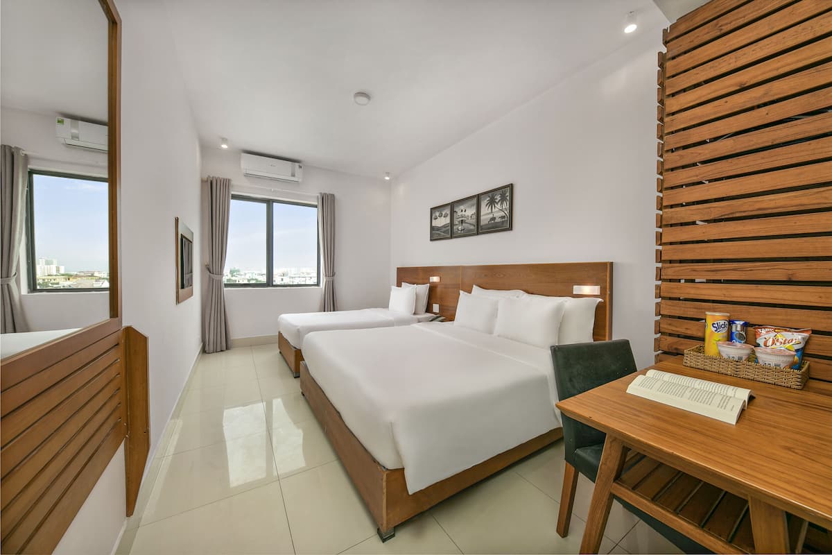 Quarto de hotel com duas camas, cabeceira de madeira, uma escrivaninha com cadeira e uma janela com cortinas.