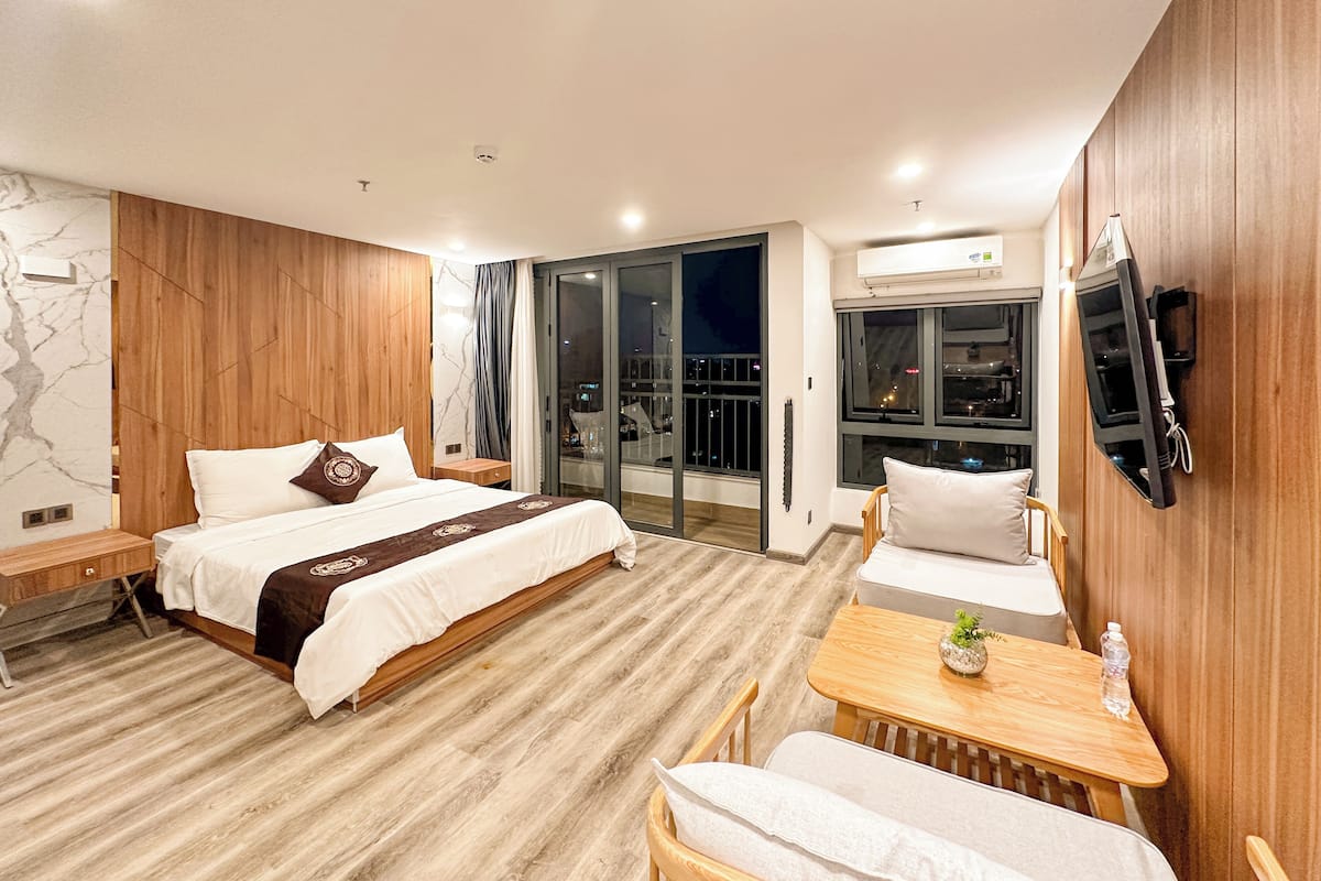 Quarto de hotel moderno com uma cama grande, uma escrivaninha de madeira, uma cadeira, uma pequena mesa com uma planta e uma TV fixada na parede.