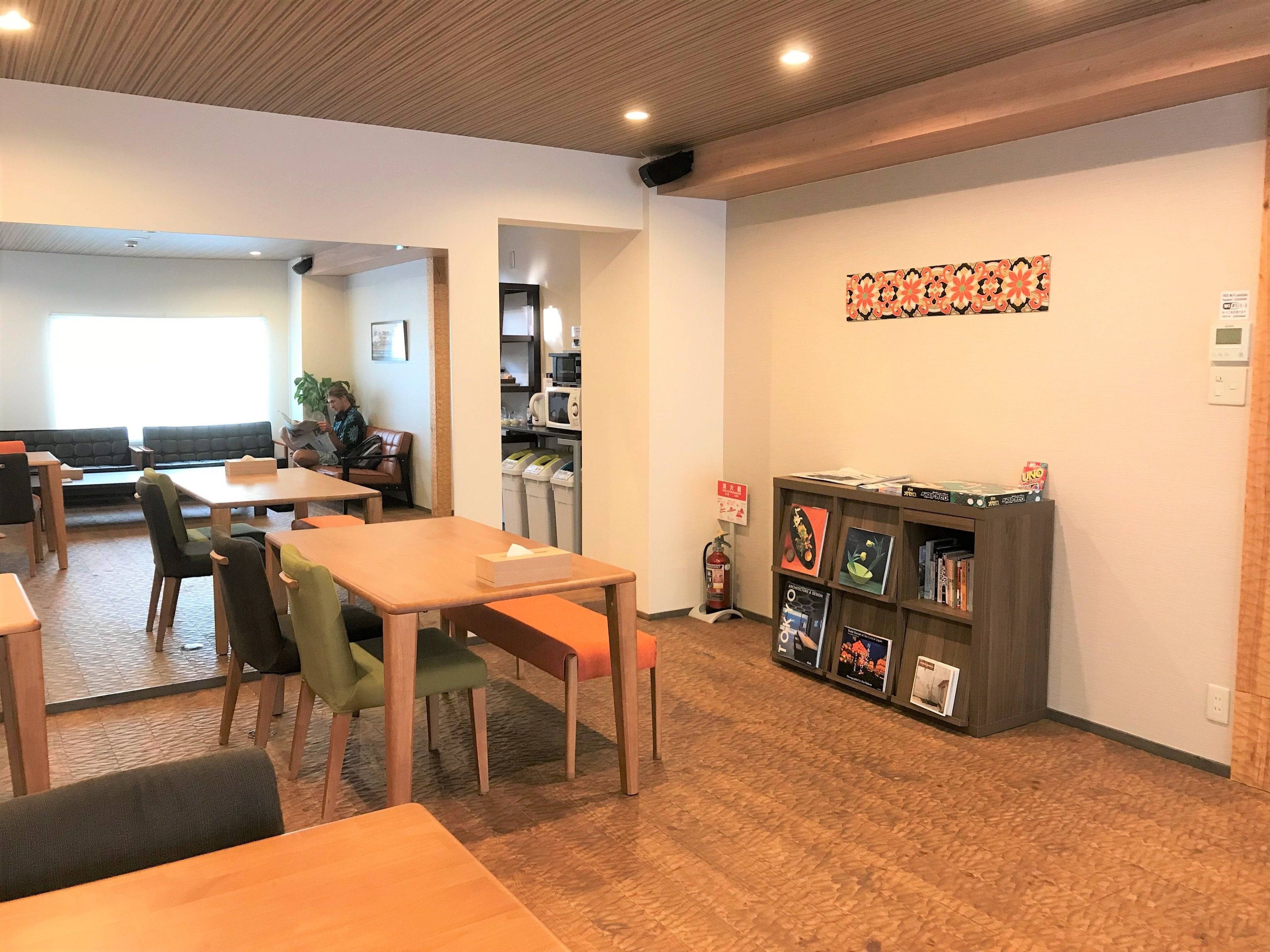 Un interior moderno con mesas y sillas de madera, una estantería con libros y una cocina al fondo.