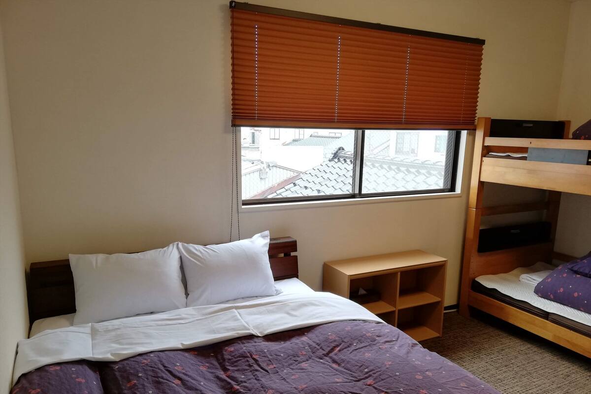 Ein Schlafzimmer mit Bett, Fenster mit Jalousien, einem Schlafsofa und einem kleinen Holztisch.