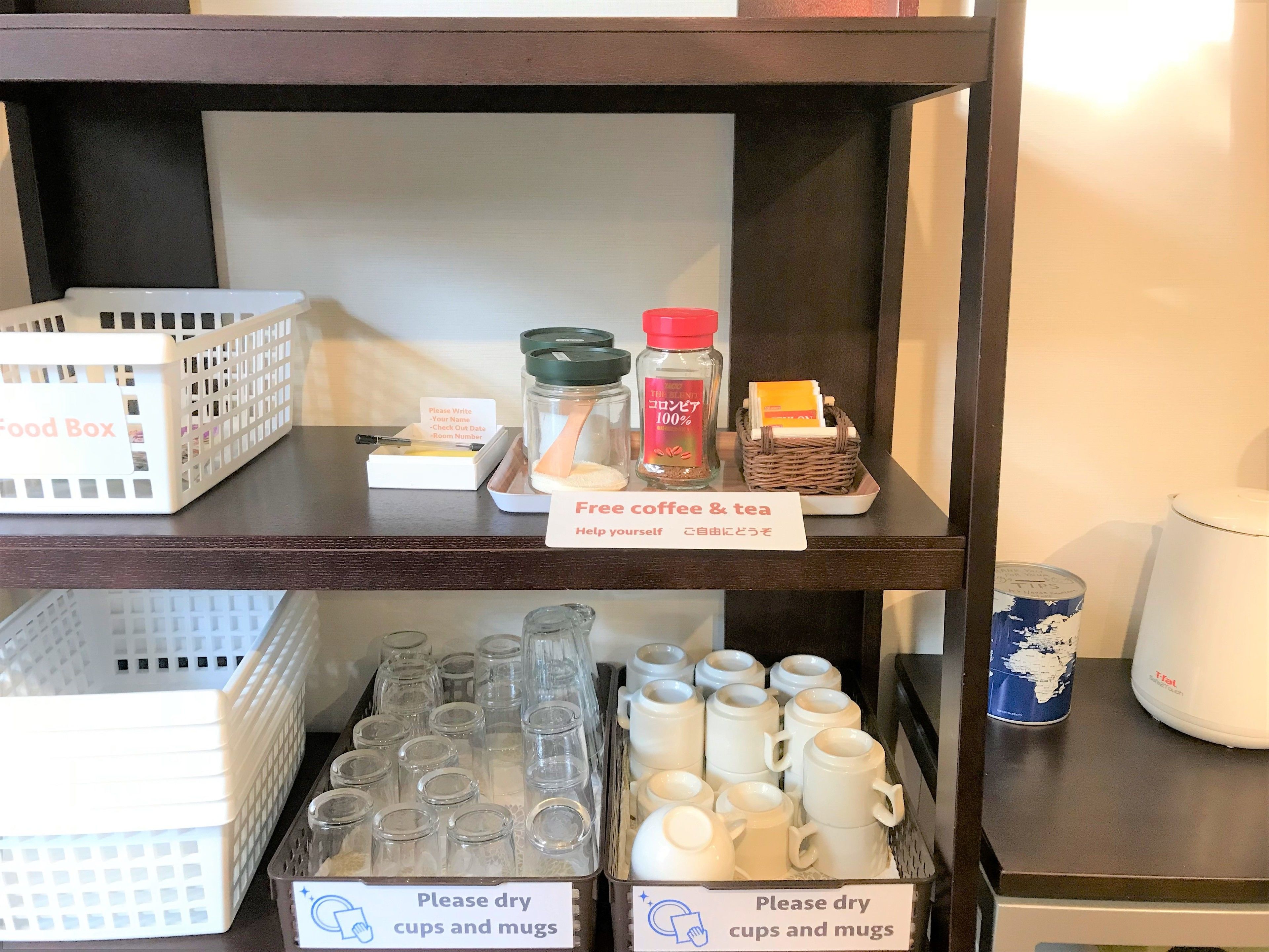 Una estantería con una caja de alimentos, un cartel que indica café y té gratis, y una bandeja con granos de café y bolsitas de té. Abajo, hay filas de tazas y boles con un letrero para secarlos.
