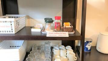 Una estantería con una caja de alimentos, un cartel que indica café y té gratis, y una bandeja con granos de café y bolsitas de té. Abajo, hay filas de tazas y boles con un letrero para secarlos.