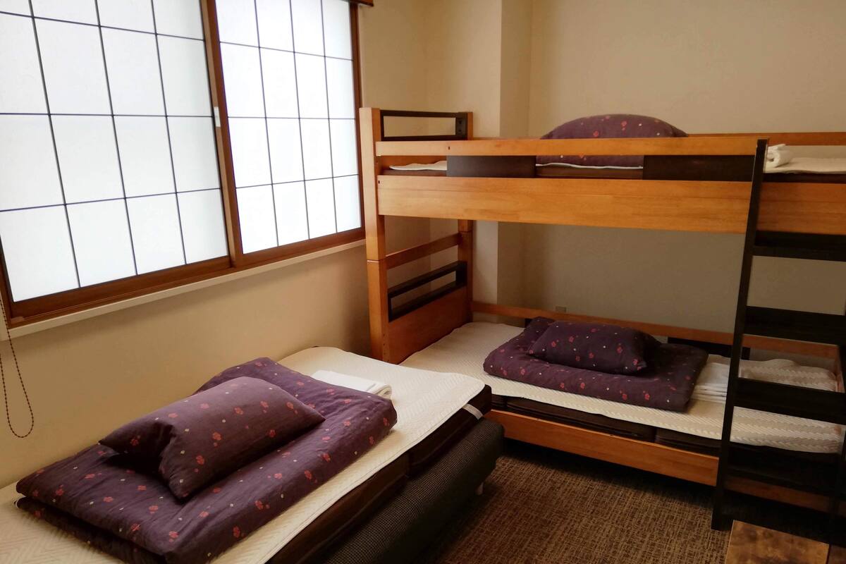 Ein Zimmer mit einem Etagenbett, einem Einzelbett mit violetten Blumenmotiven und einem Fenster mit Gittervorhängen.