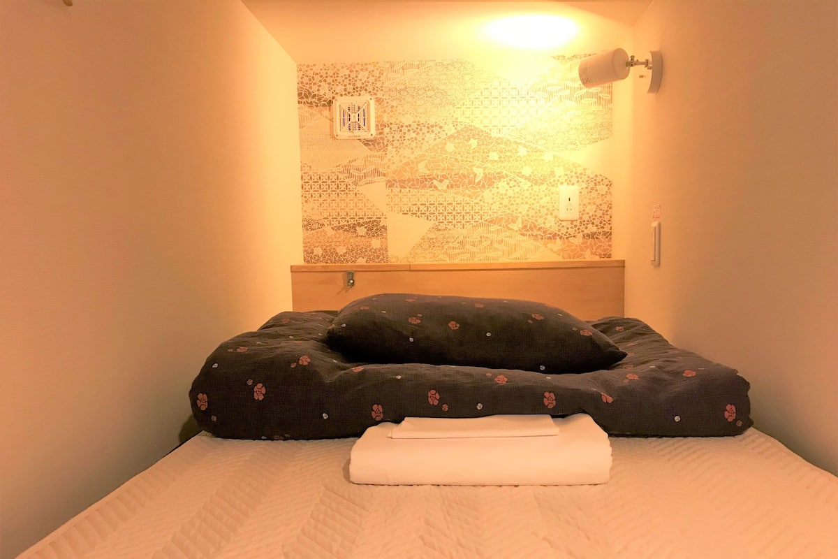 Ein ordentlich bezogenes Bett mit weißer Decke und einem blumigen Kissen, einer Wandleuchte und einem dekorativen Wandmuster.