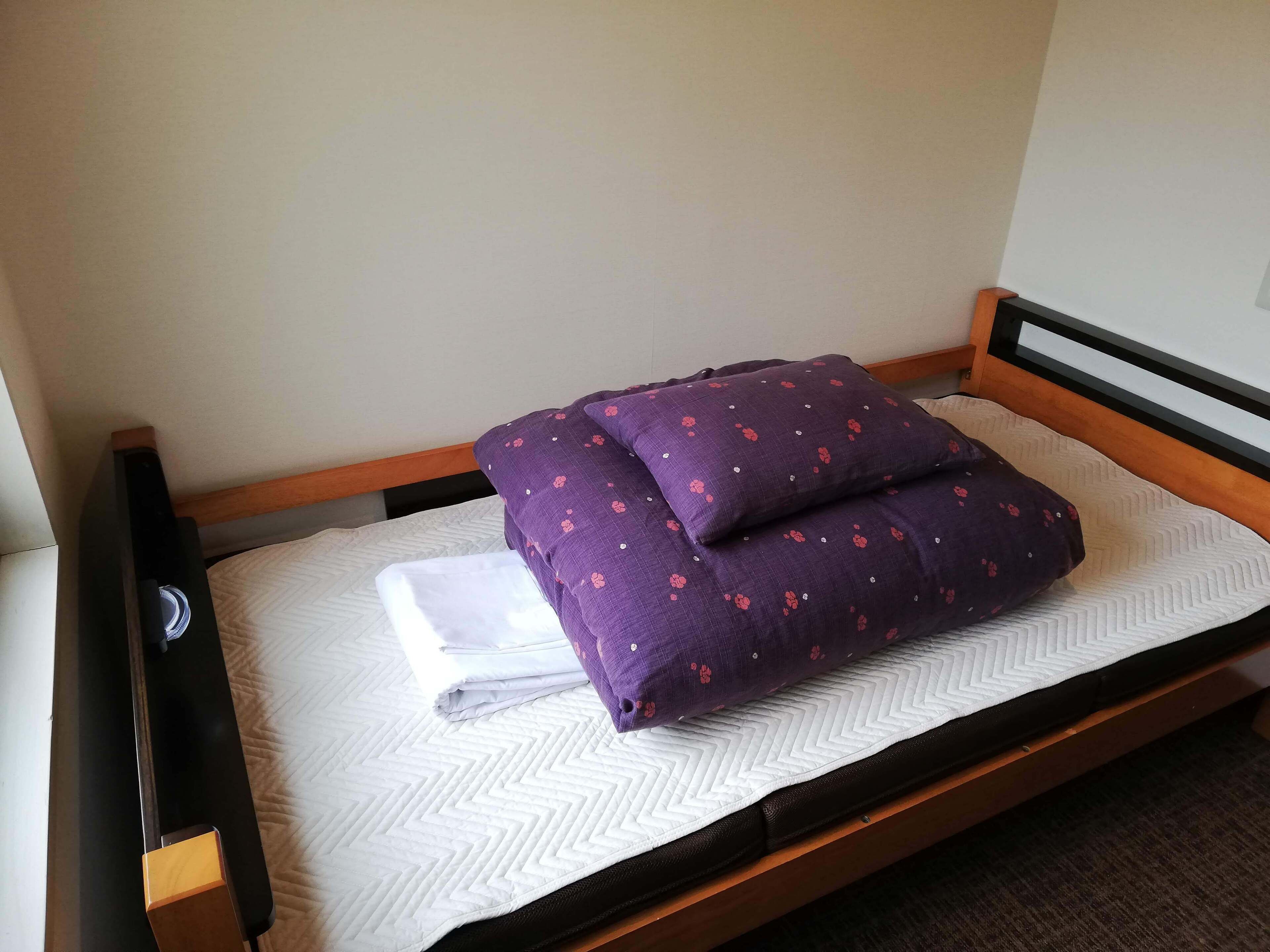 A single bed with a wooden frame, a white quilt, and two purple pillows with a floral pattern.