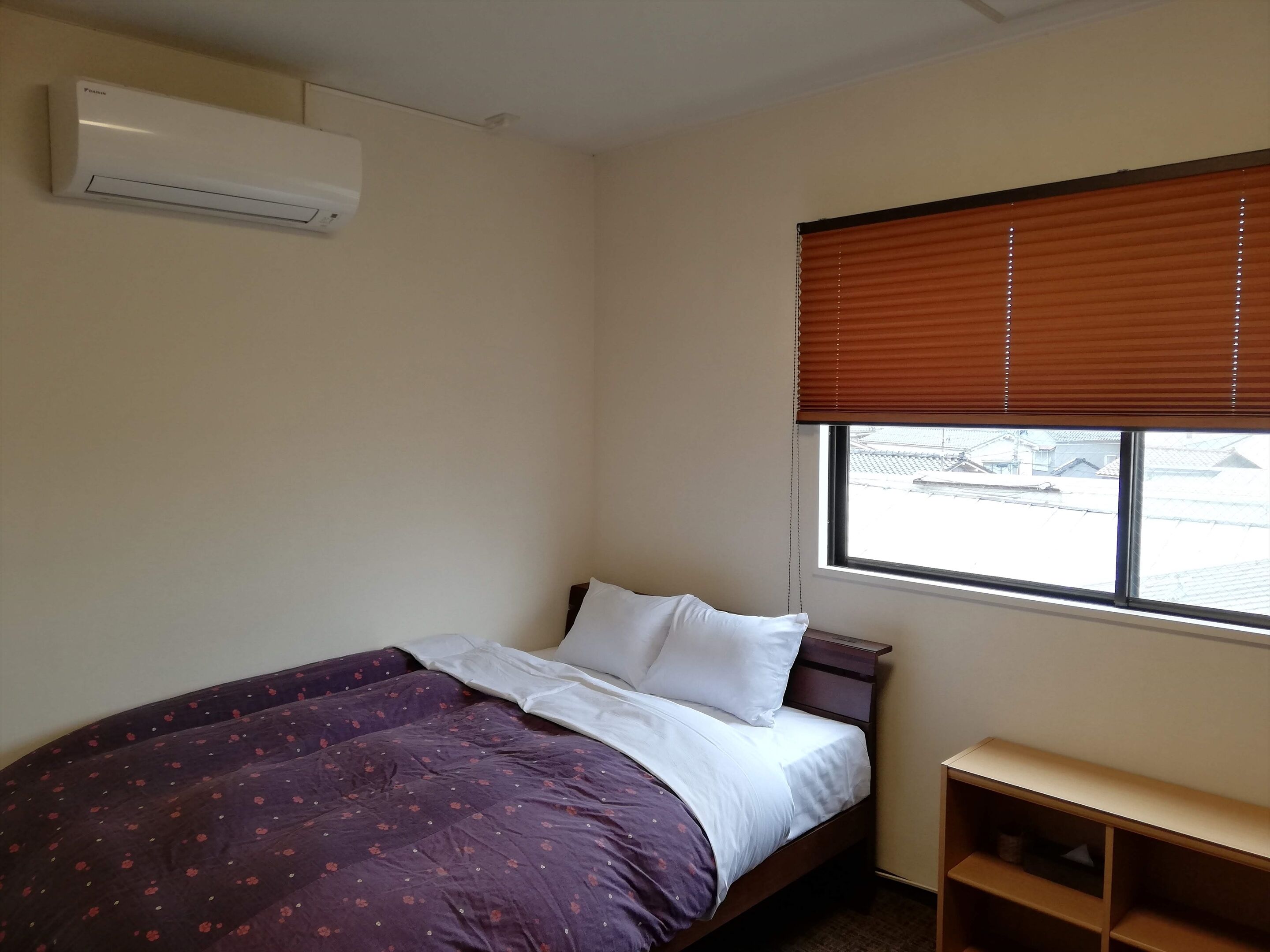 A bedroom with a bed, a window with blinds, a bedside table, and an air conditioning unit.