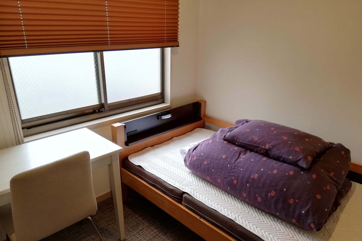 Ein kleines Zimmer mit einem Einzelbett, einem Schreibtisch und einem Stuhl, einem Fenster mit Jalousien und einem Bett mit violett gemusterten Blumenbezügen.