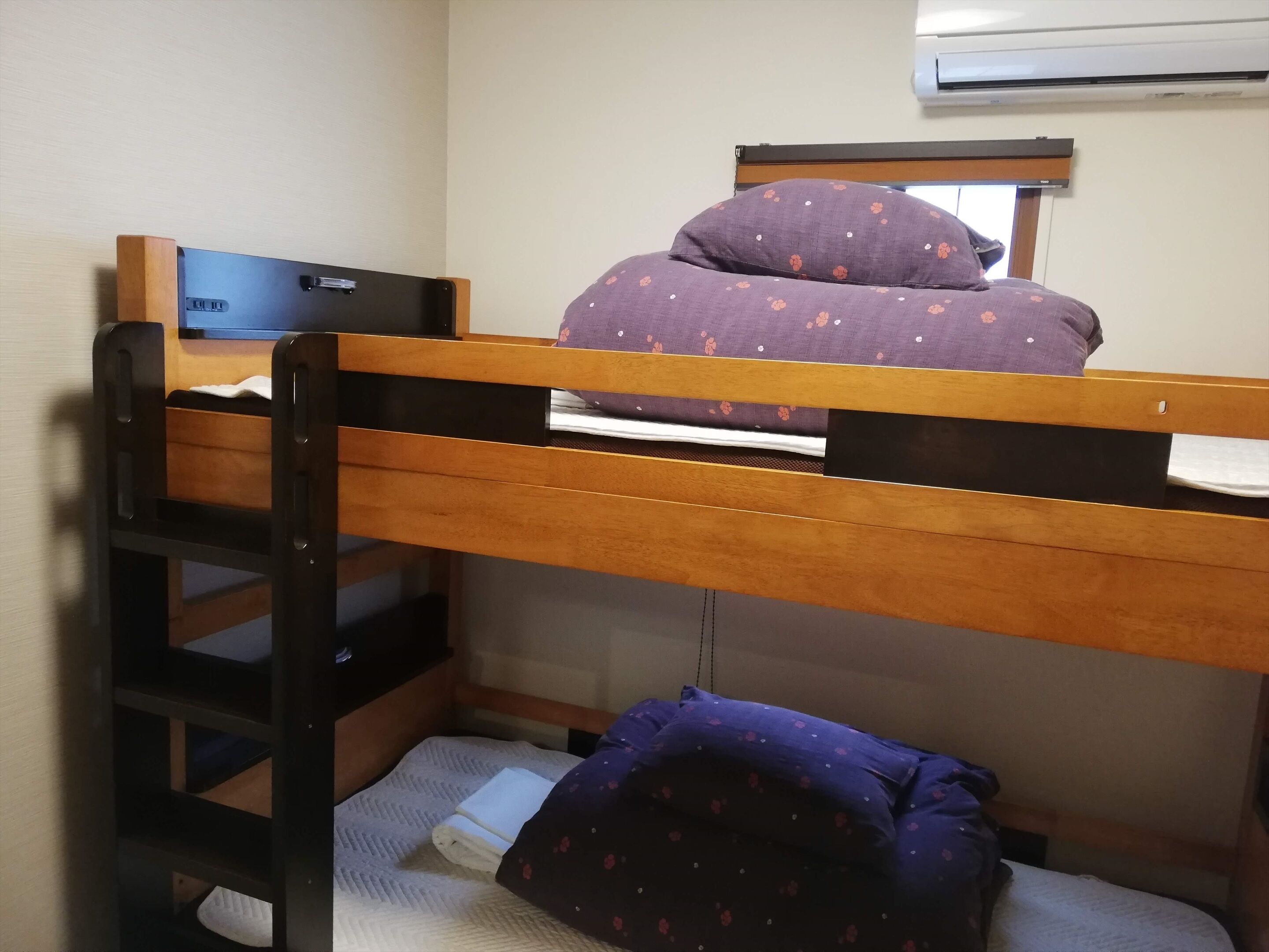 A bunk bed with a laptop on the top bunk, a window with a blind, and an air conditioning unit above.