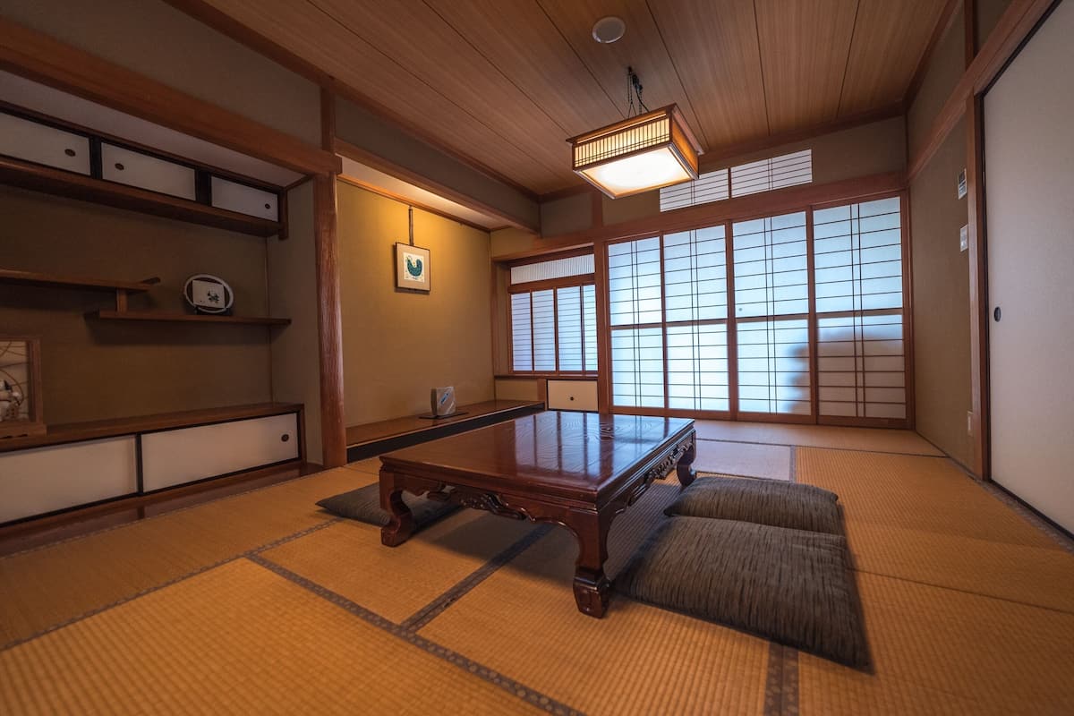 Ein traditioneller japanischer Raum mit Tatami-Bodenbelag, einem niedrigen Holztisch, verschiebbaren Shoji-Türen und einer Deckenleuchte.