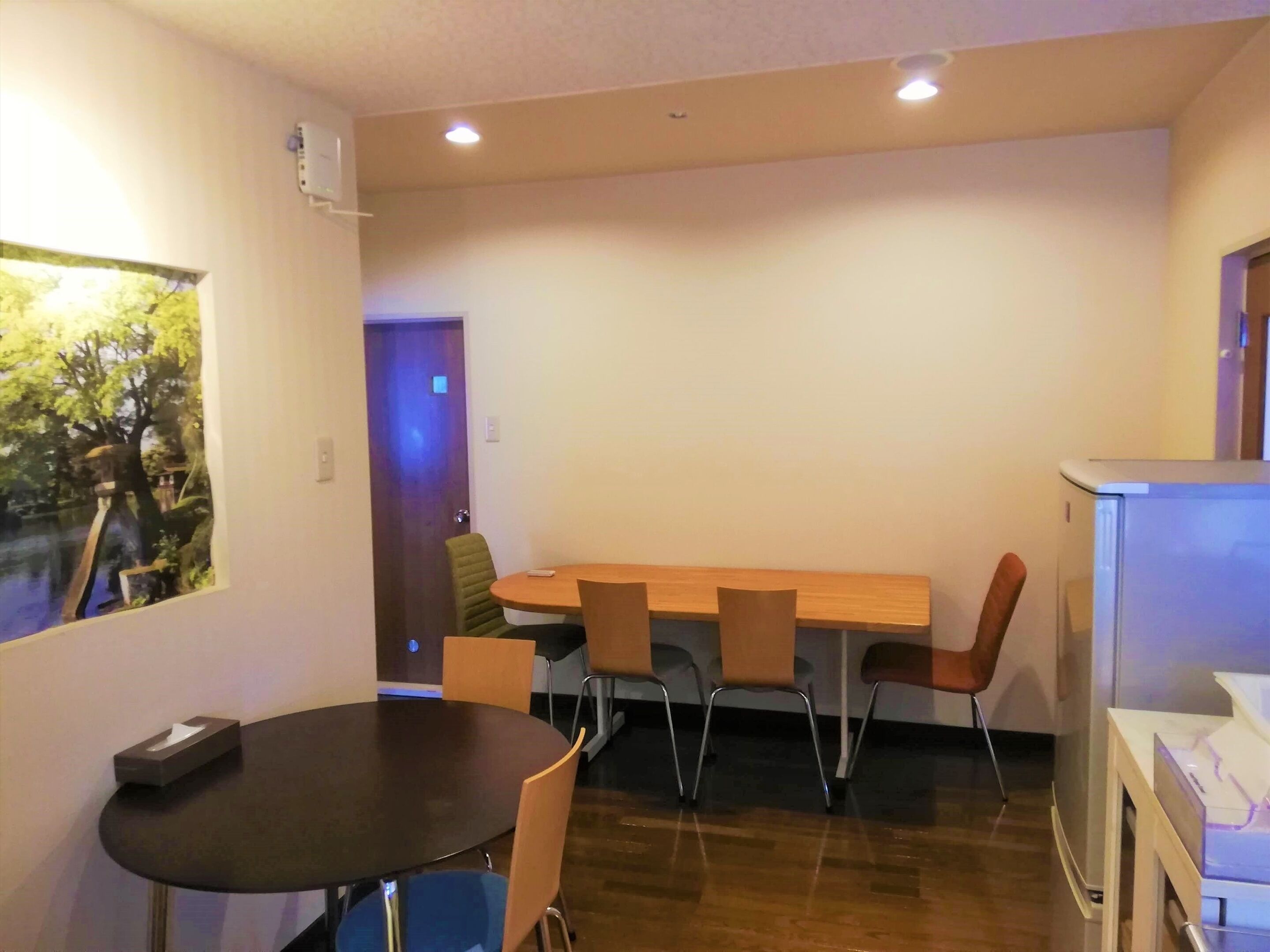A room with a wooden dining table, four chairs, a small round table with a tissue box, and a refrigerator.