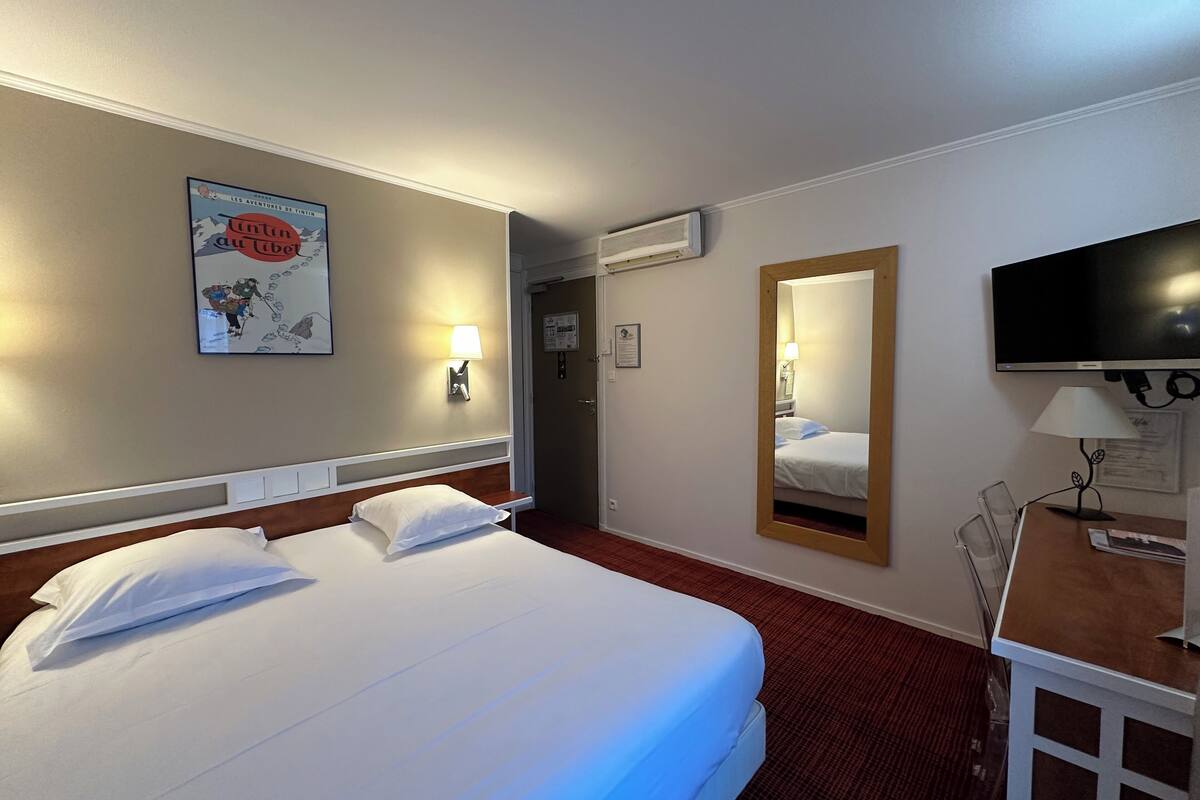 A hotel room with a large bed, a desk with a chair, a TV, a mirror, and a wall-mounted lamp.