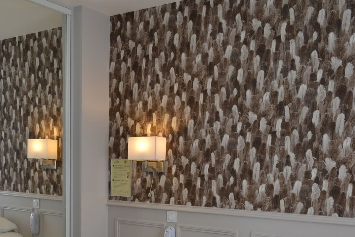 A hotel room with a large bed, two red pillows, a nightstand with a lamp, a mirror, and a wall with a feather pattern.