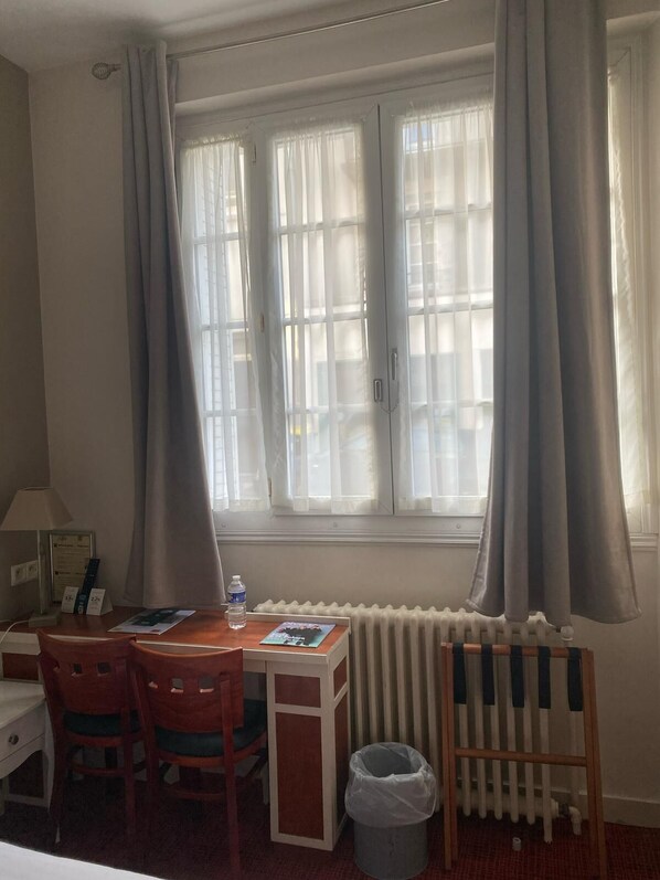 Quarto de hotel com escrivaninha, cadeiras, radiador e janela com cortinas.
