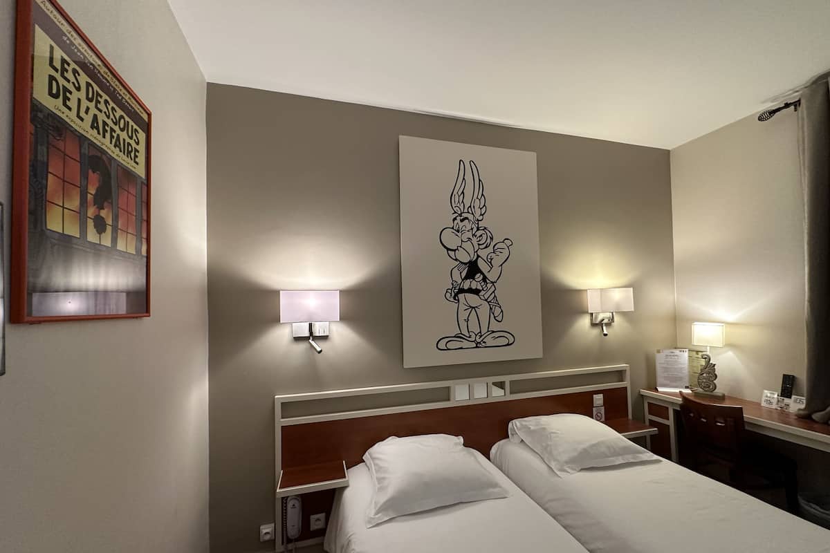 A hotel room with two beds, a headboard, bedside lamps, a wall-mounted artwork of a cartoon character, and a framed picture on the wall.