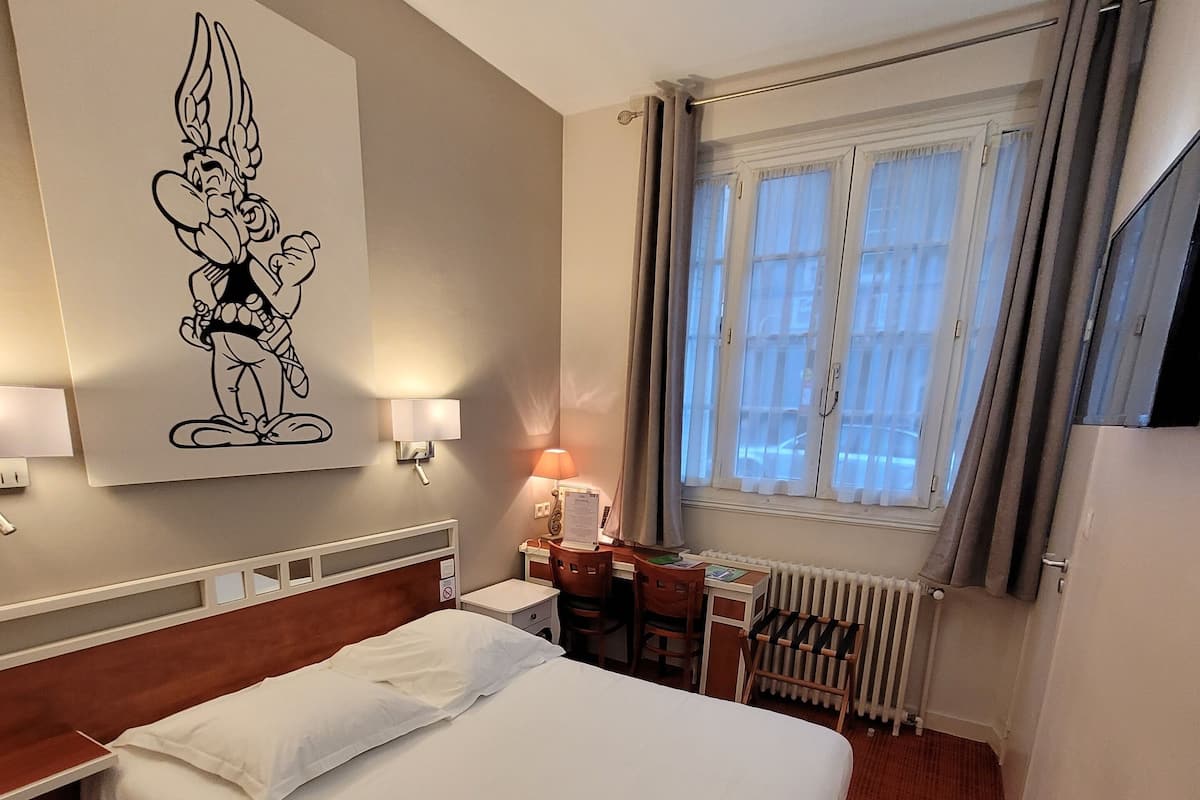 A hotel room with a large bed, a desk with a chair, a radiator, a window with curtains, and a wall-mounted TV.