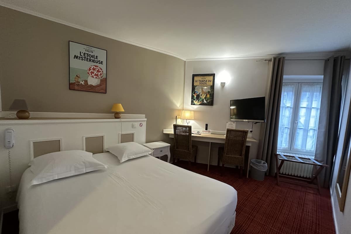 A hotel room with two beds, a desk, a chair, a TV, a lamp, and a poster on the wall.