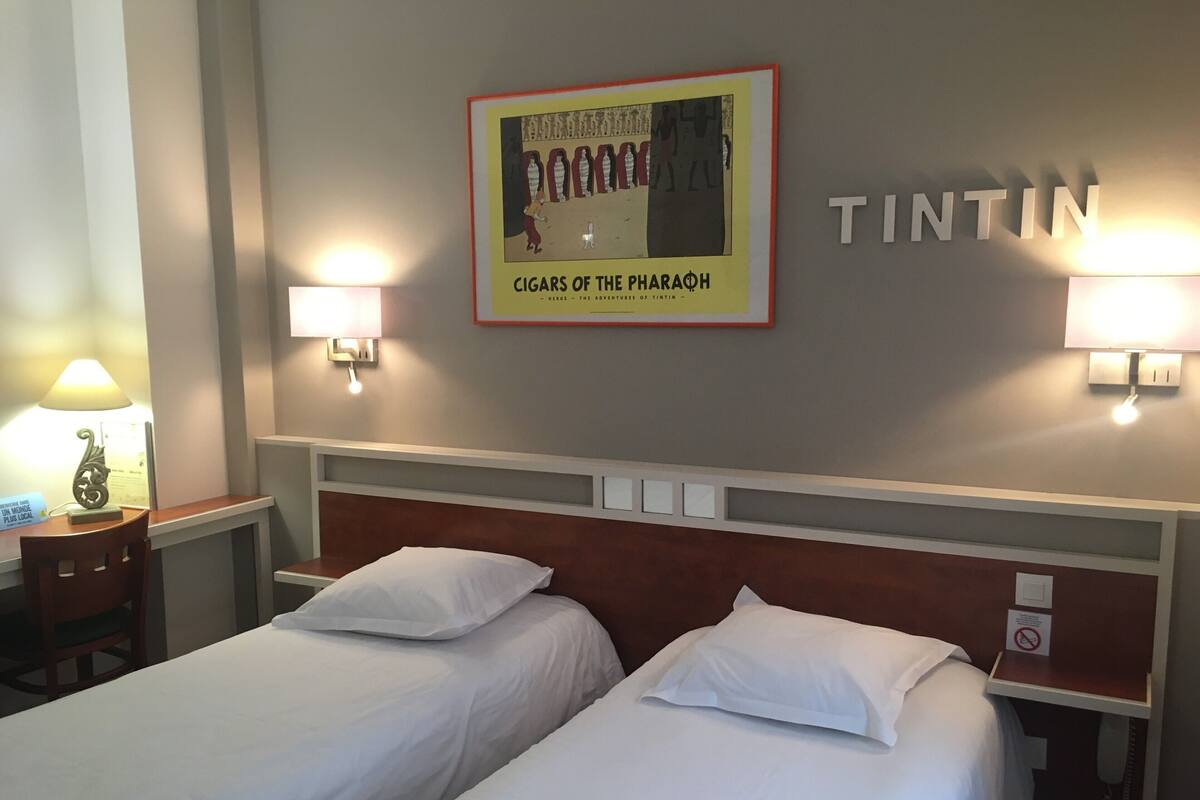A hotel room with two beds, a framed poster on the wall, and two wall-mounted lamps.