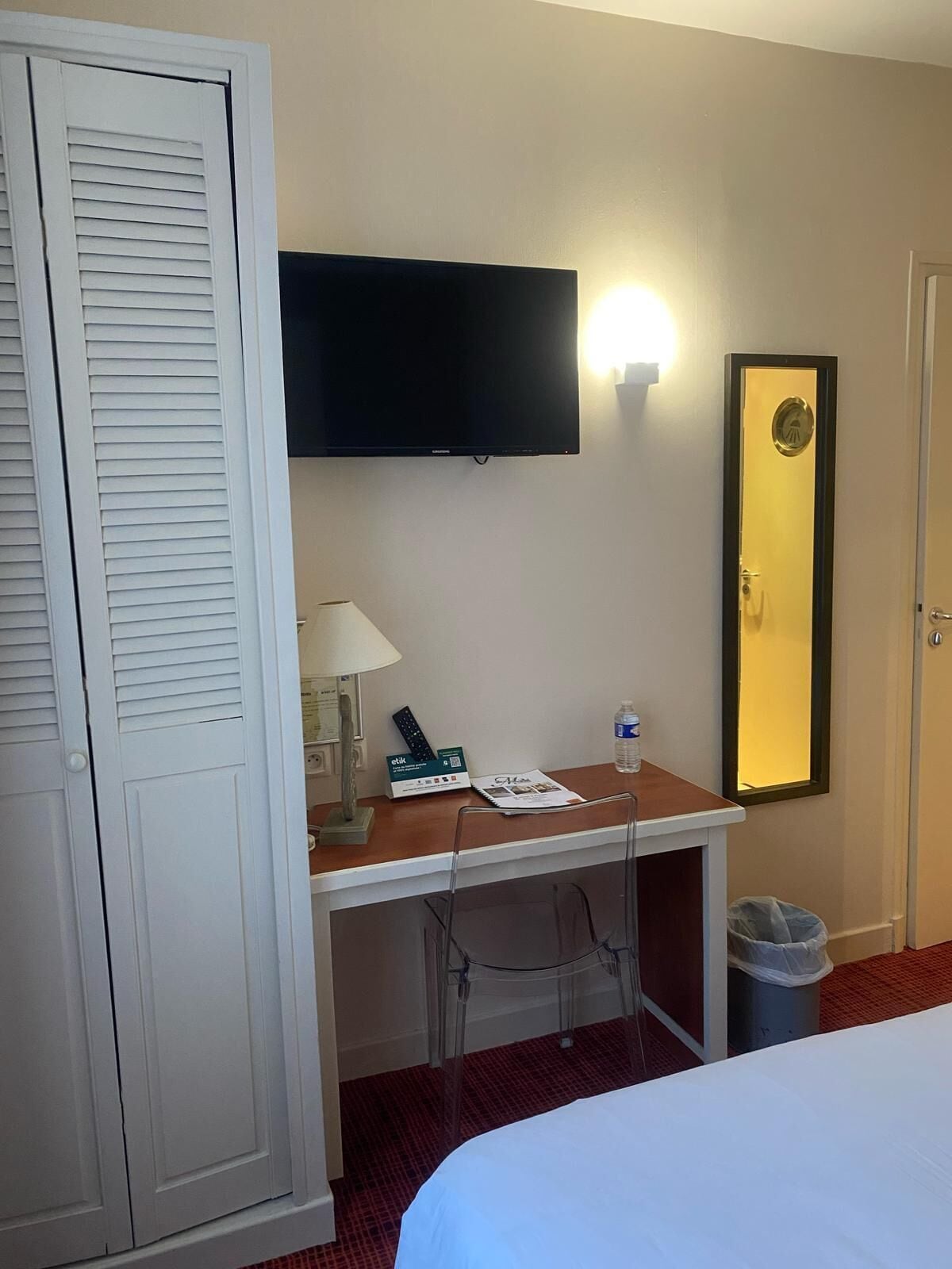 Una habitación de hotel con cama, escritorio con lámpara, televisión y espejo.