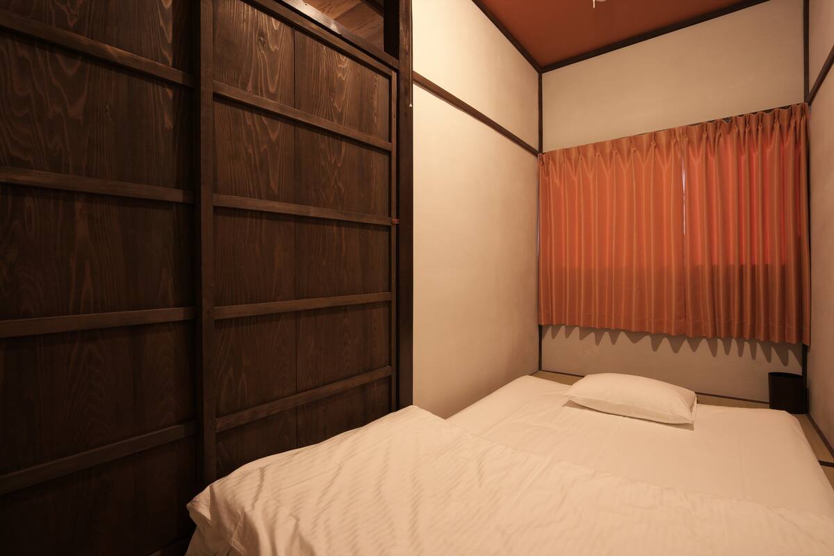 A bedroom with a wooden wardrobe, a bed with white bedding, and a window with orange curtains.