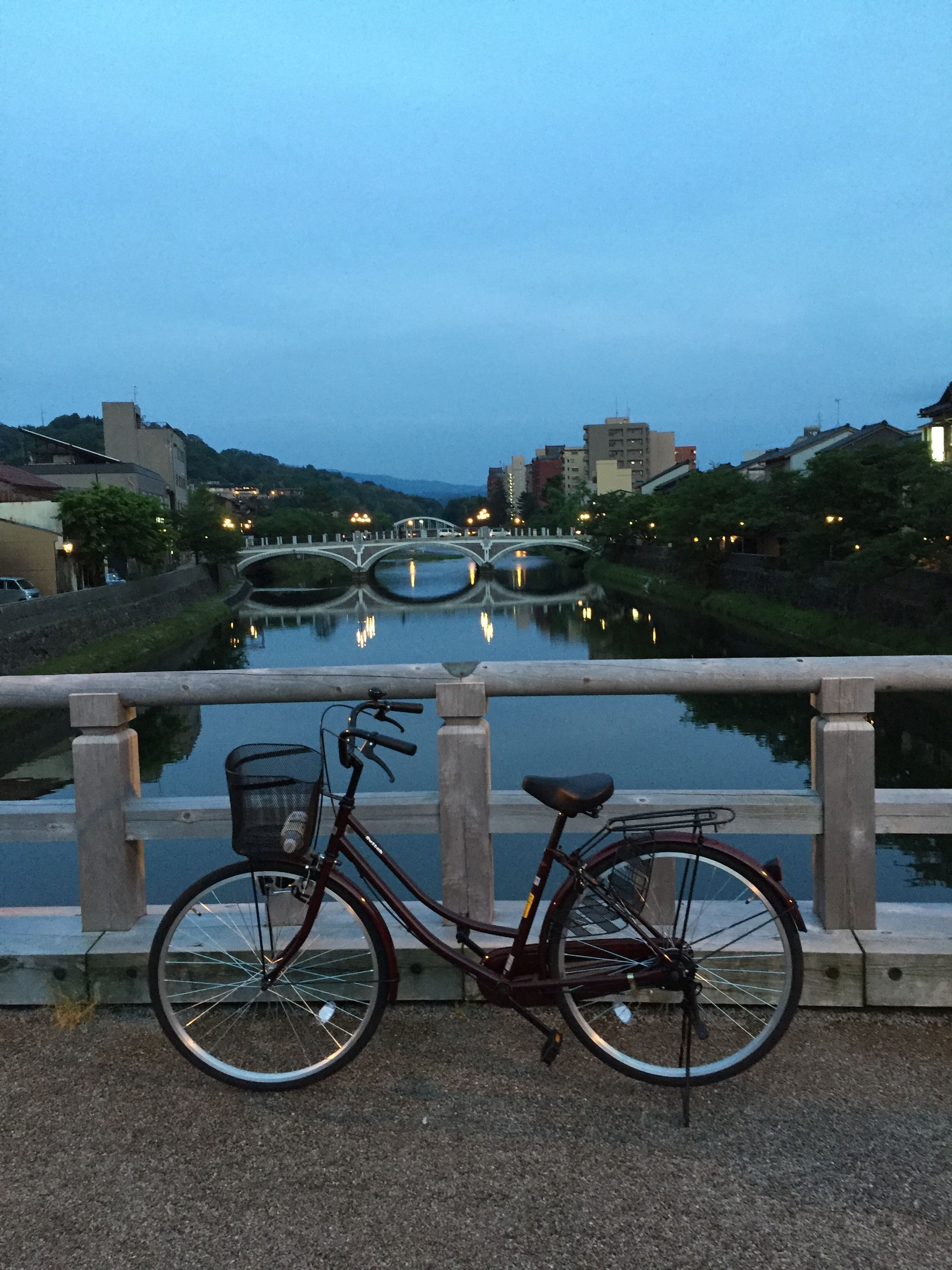A bicycle parked by a calm river with a bridge in the background.