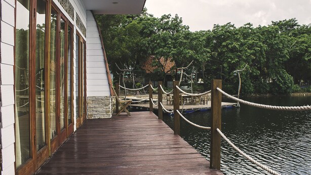 A wooden dock extending over water, with a building on the left featuring large windows and a stone base. - Kanaka Lake Villa (Batam)