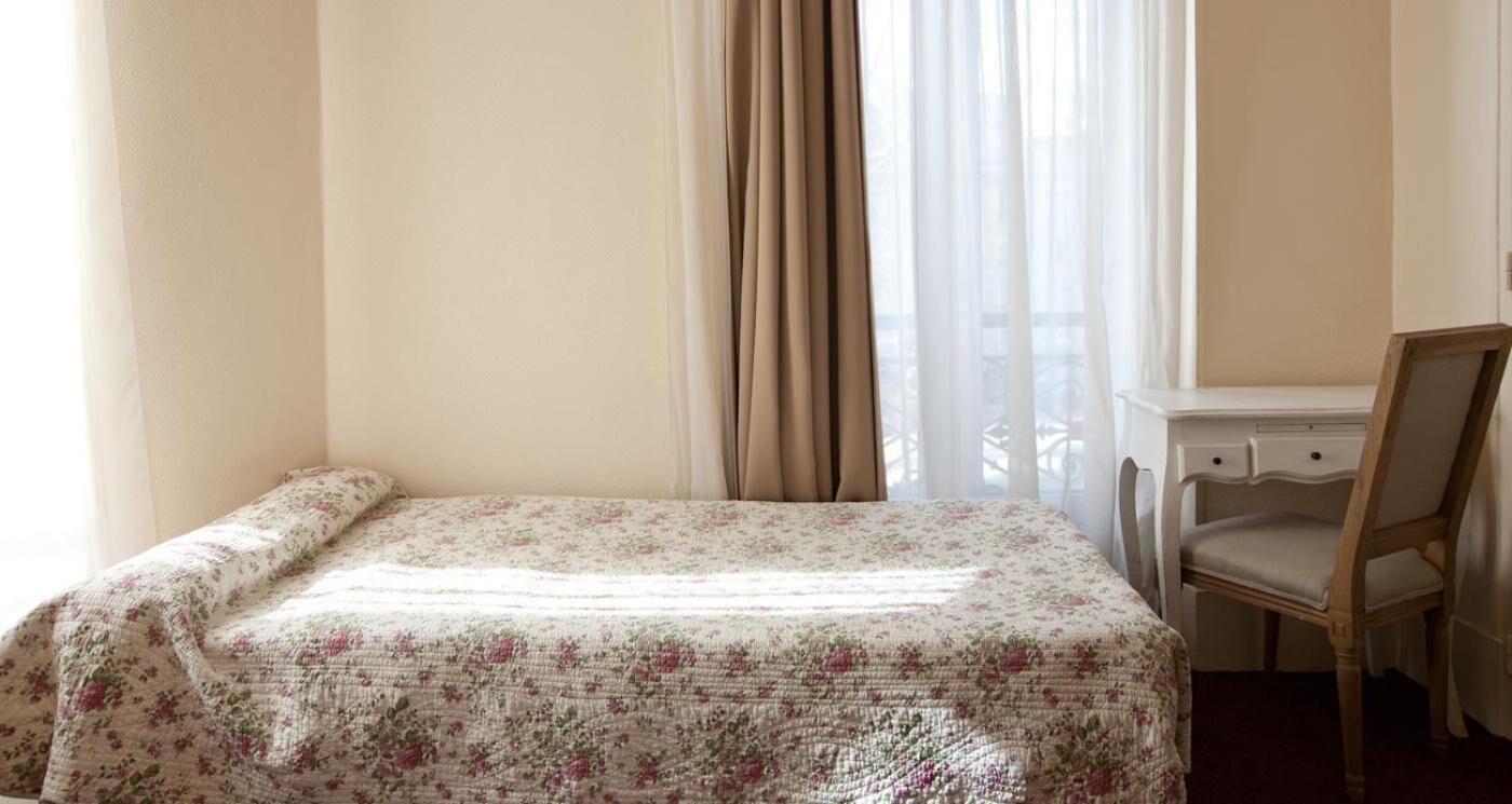 A room with a bed covered in a floral bedspread, a chair, and a desk with a chair.