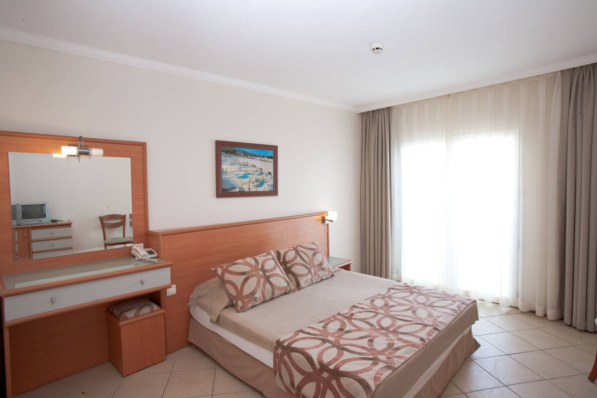 Una habitación de hotel con cama, escritorio con teléfono, silla, espejo y un cuadro en la pared.