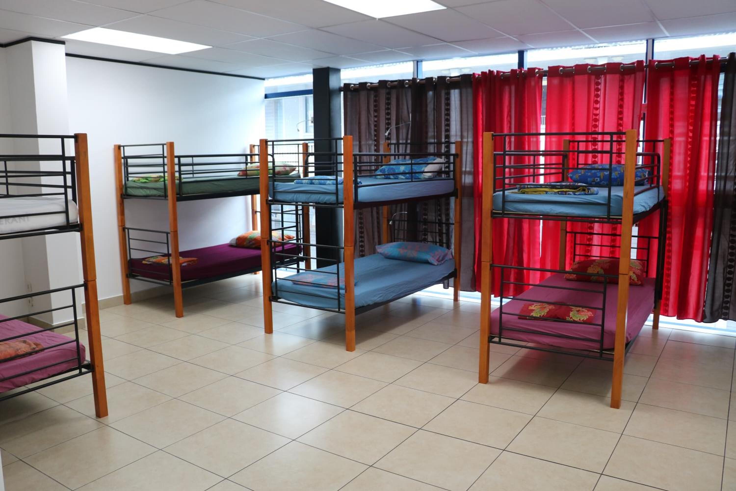 A dormitory room with bunk beds, a window with red curtains, and a tiled floor.