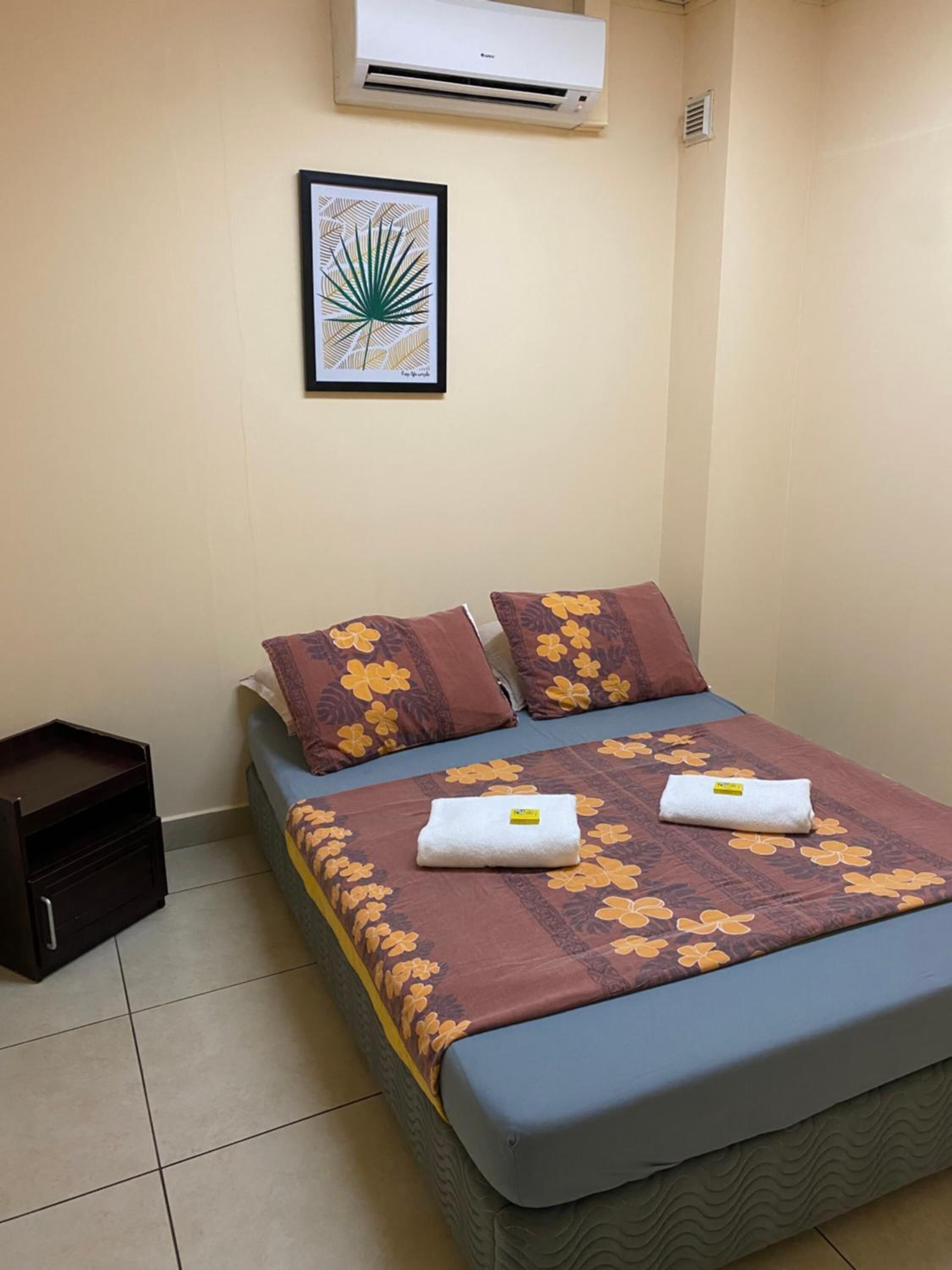A single bed with floral bedding, two pillows, a small bedside table, and an air conditioning unit on the wall.