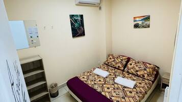 A hotel room with a bed, a small table, a trash bin, and a wall-mounted air conditioner.