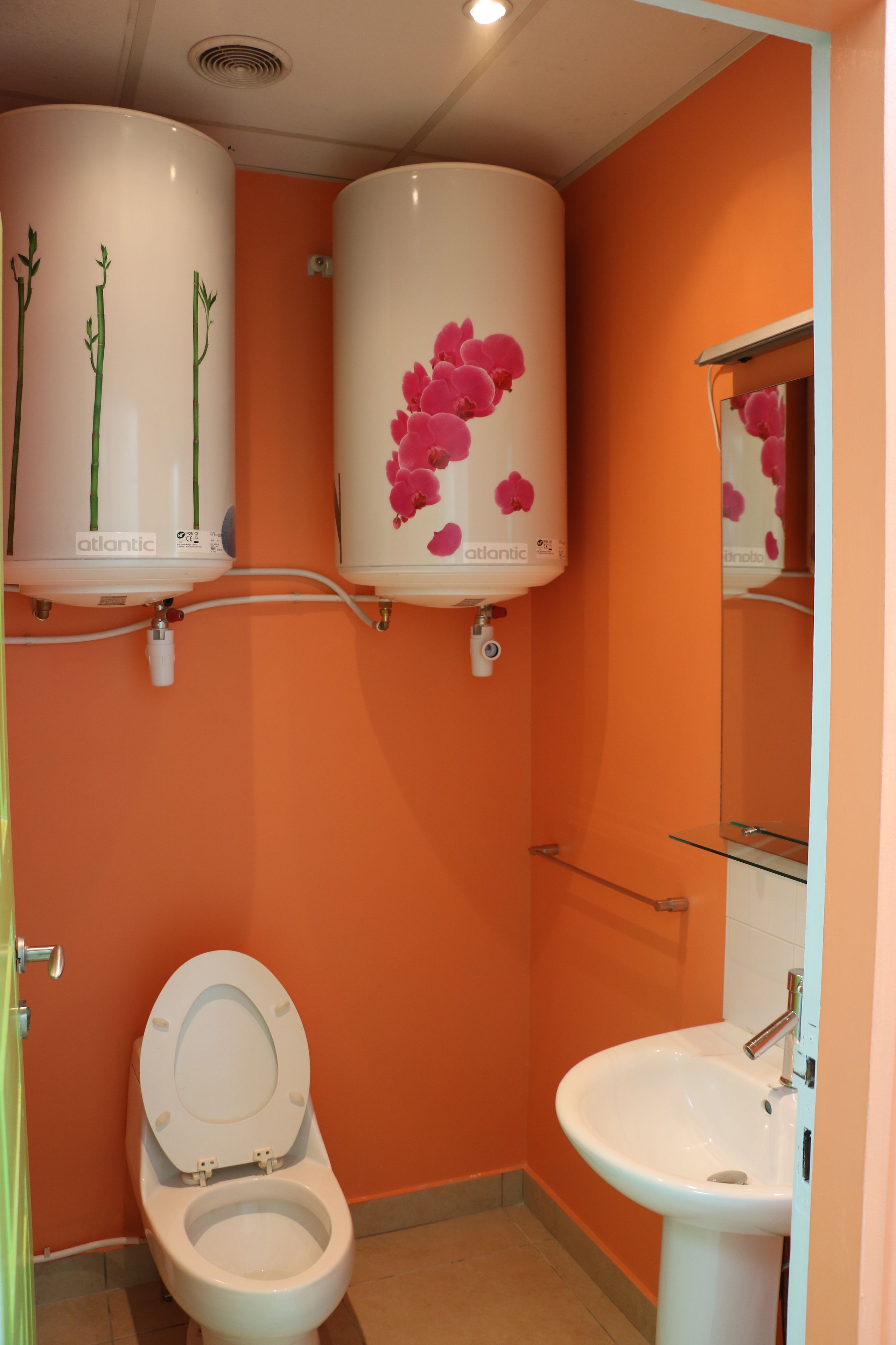 Un baño con pared naranja, inodoro blanco, lavabo blanco y dos calentadores de agua blancos con diseños florales.