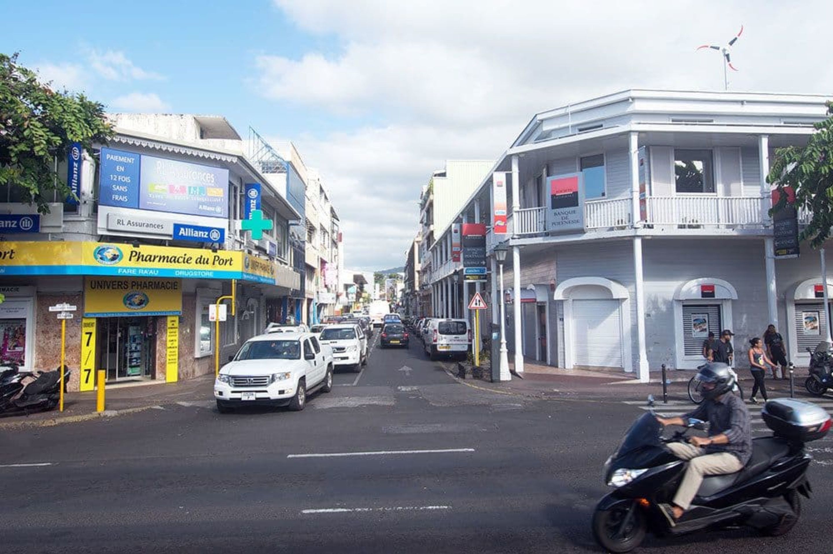 Straßenansicht mit einer Apotheke, einem Apothekenzeichen und einem weißen Auto, das am Straßenrand geparkt ist.