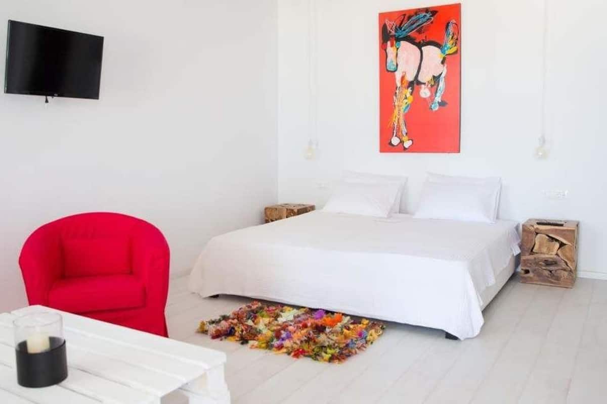 Un dormitorio con una cama, una silla roja, una mesa blanca, un televisor en la pared y una obra de arte colorida.