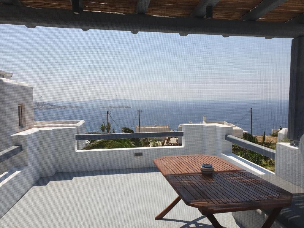 A balcony with a wooden table and chairs, overlooking a coastal landscape with buildings and the sea.