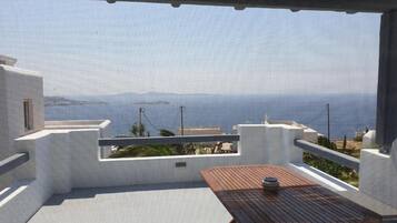 A balcony with a wooden table and chairs, overlooking a coastal landscape with buildings and the sea.