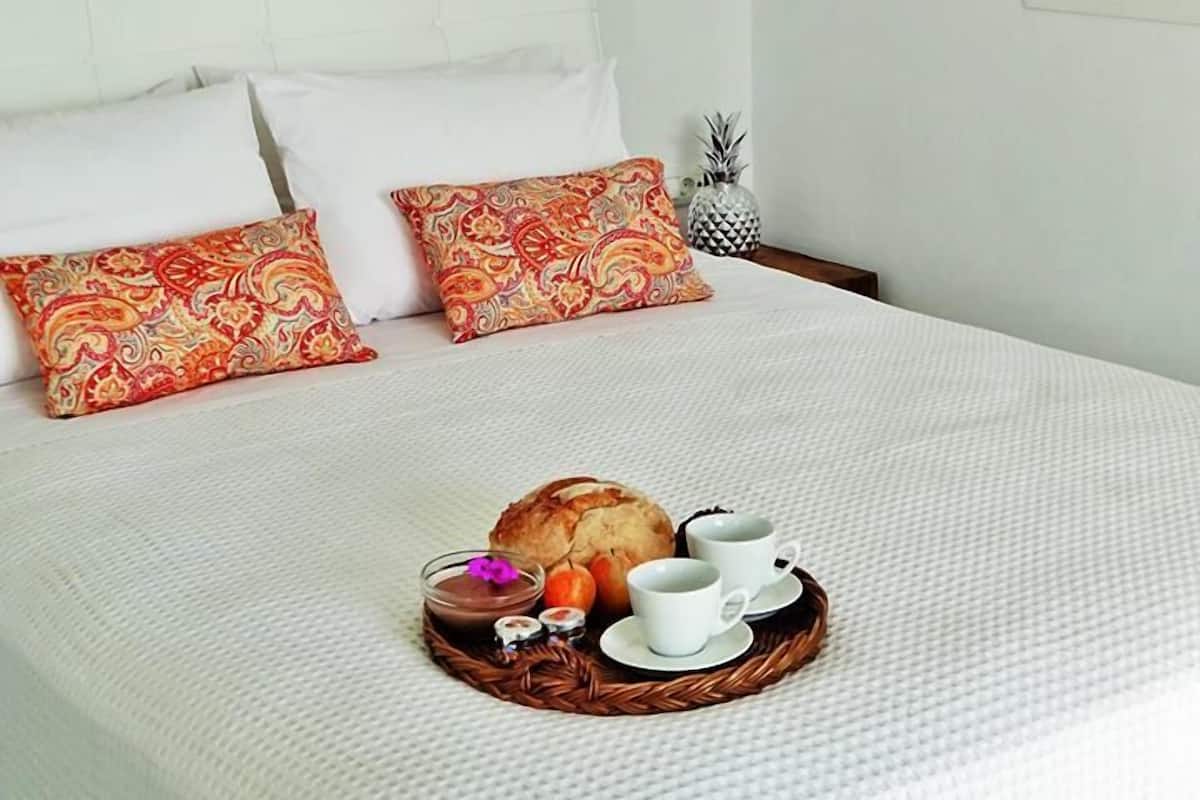 Una cama bien hecha con sábanas blancas y almohadas estampadas, una bandeja con artículos de desayuno y una lámpara colgante.