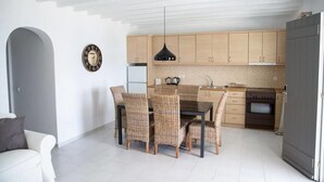 A modern kitchen with light wood cabinets, a black pendant light, and a dining area with wicker chairs. - Bellou Suites (Mykonos)