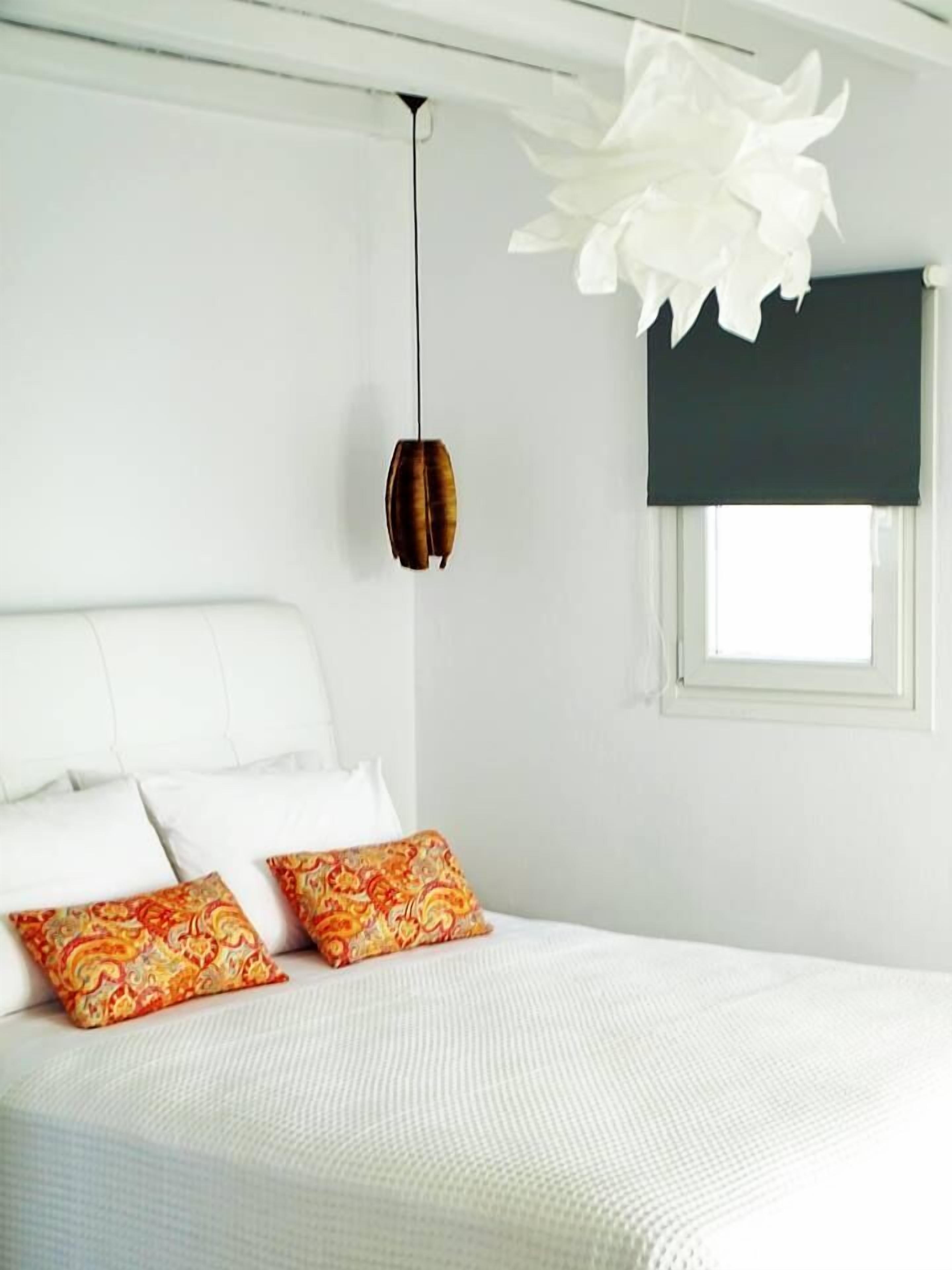 A bedroom with a white bed, two patterned pillows, a hanging light fixture, and a window with a dark blind.