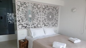 A neatly made bed with white linens and two rolled towels, a decorative headboard, and a small wooden side table. - Bellou Suites (Mykonos)