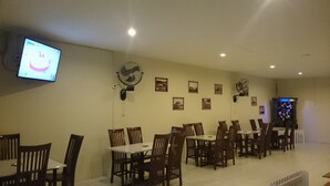 A dining area with multiple tables and chairs, a television mounted on the wall, and several wall-mounted fans. - De Ritz Hotel (Surabaya)