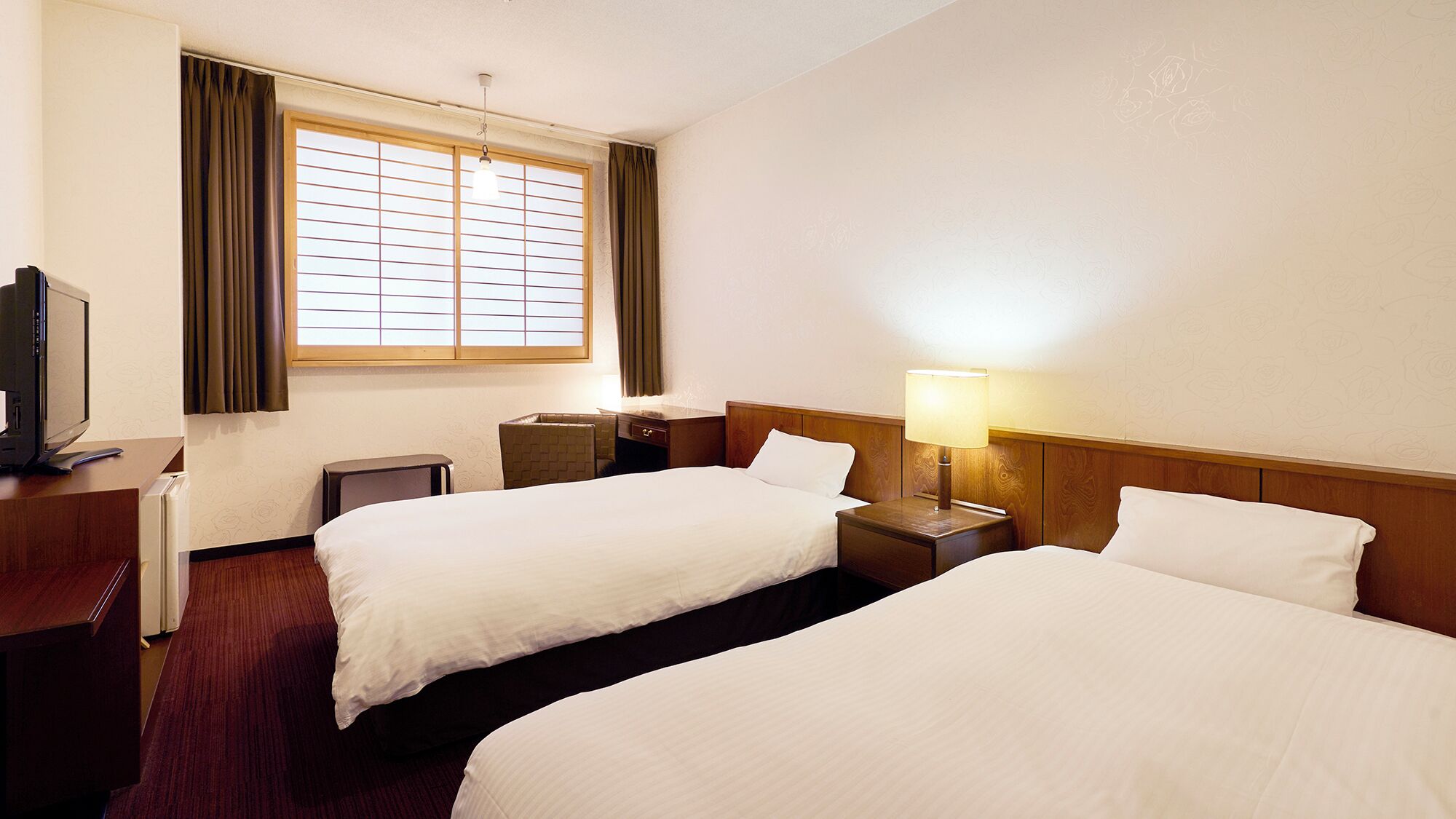 A hotel room with two beds, a desk with a computer, a lamp, and a window with curtains.