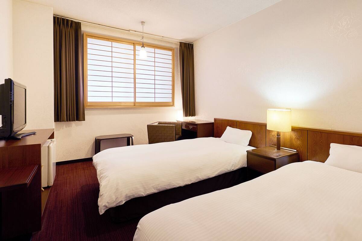 A hotel room with two beds, a desk with a computer, a lamp, and a window with curtains.