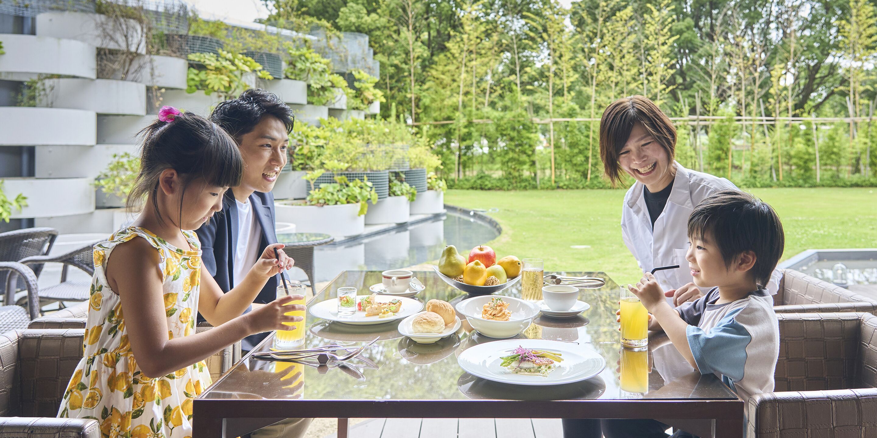 A family dining outdoors with a table set for a meal, surrounded by greenery and a modern building.