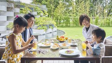 A family dining outdoors with a table set for a meal, surrounded by greenery and a modern building.
