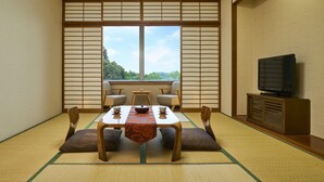 A traditional Japanese room with tatami flooring, a low table with cushions, a television, and a view of greenery outside.