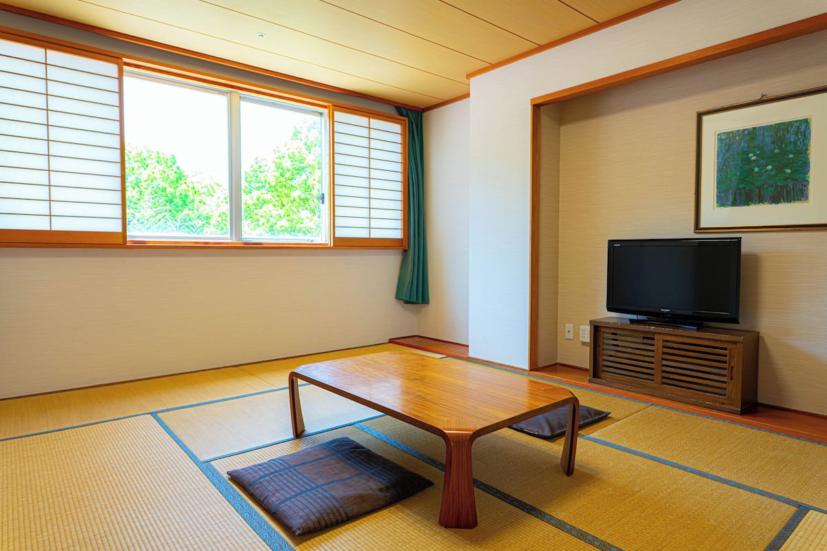 A traditional Japanese room with tatami flooring, a low wooden table, a television on a wooden stand, and a window with shoji screens.