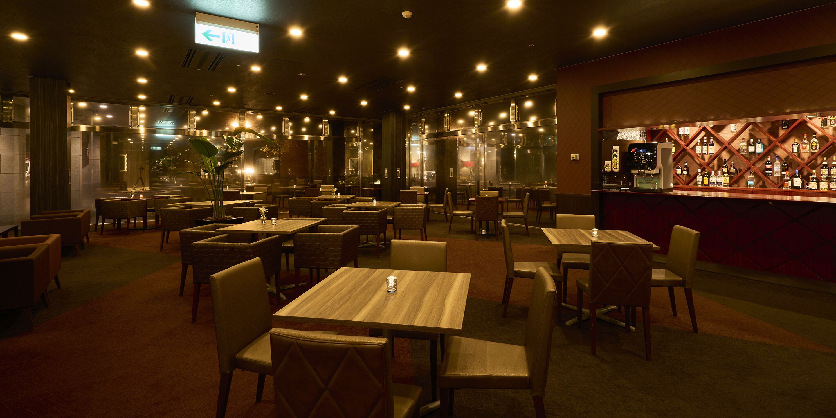 A restaurant with multiple tables and chairs, a bar area with bottles, and a large window in the background.