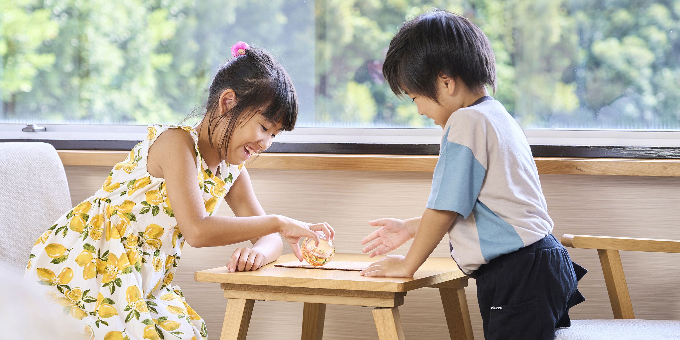 Two children playing with a ball on a wooden table in a room with a window and a couch.