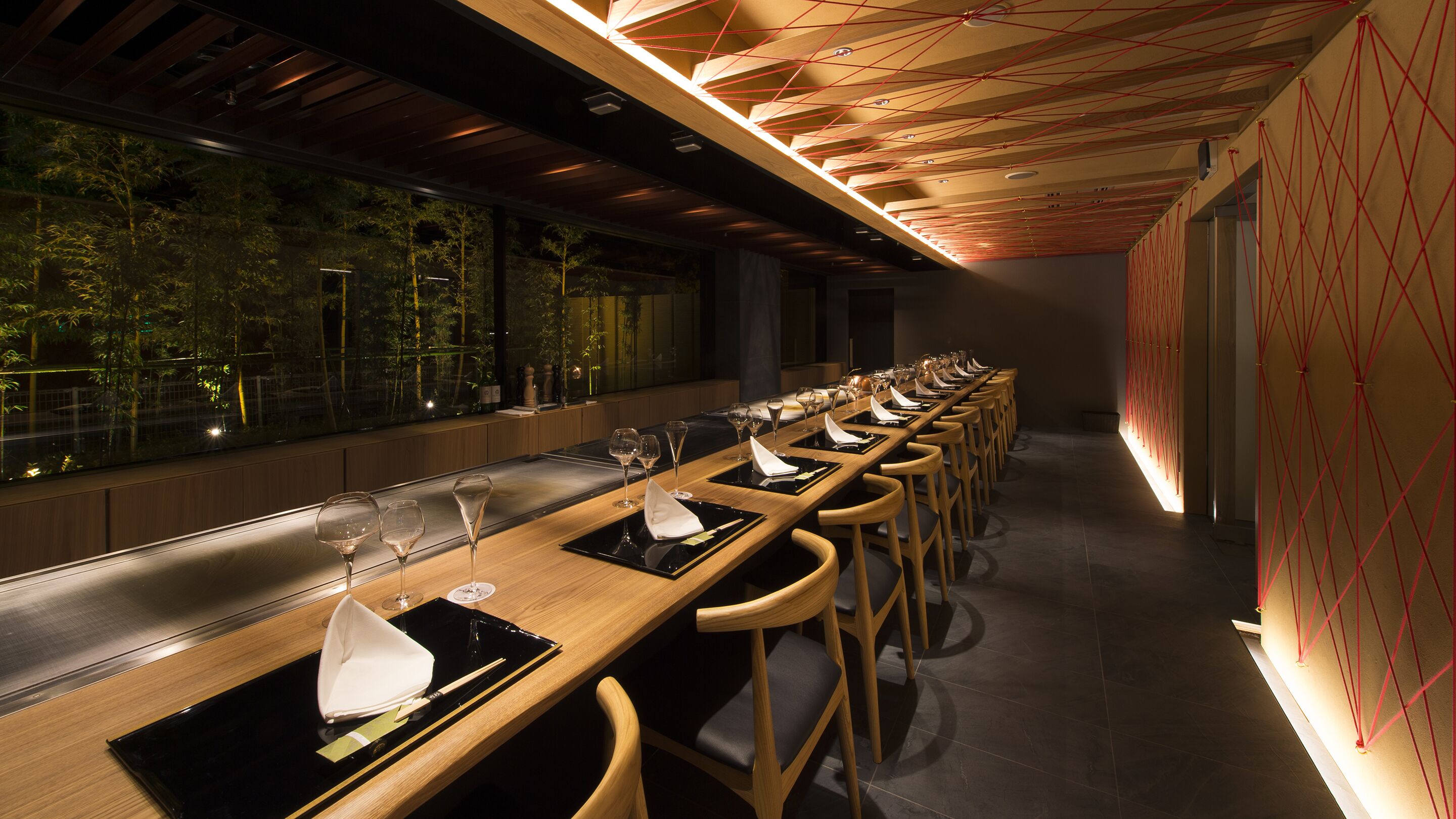 Een lange eettafel met stoelen, gedekt met wijnglazen en servetten, in een modern interieur met een houten plafond en uitzicht op bomen.