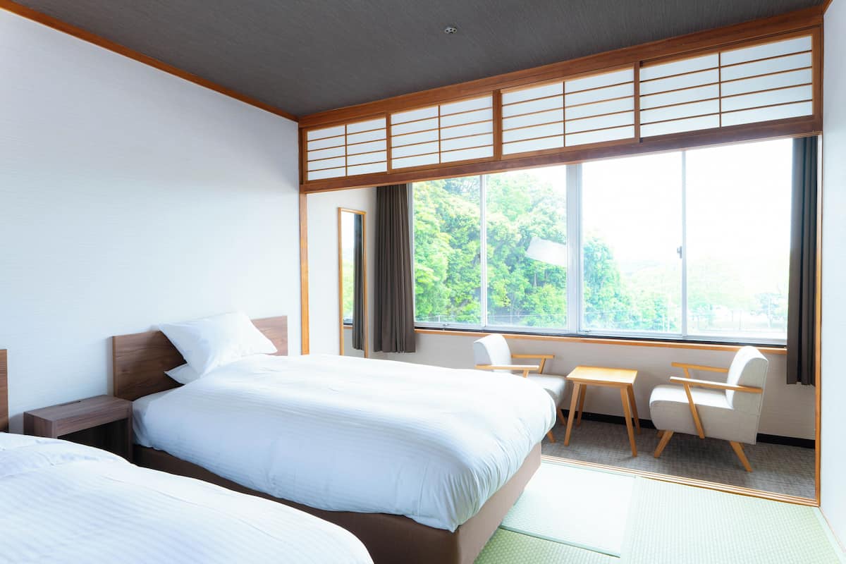 A hotel room with two beds, a desk, a chair, and a large window with a view of trees.